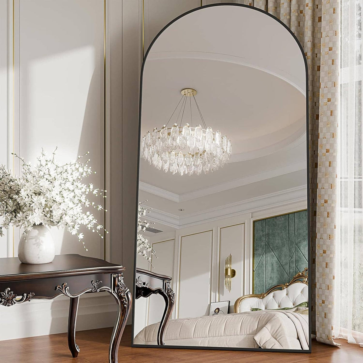 Arched full-length floor mirror with black frame, reflecting a chandelier and bed area.