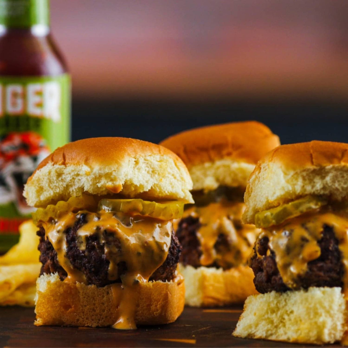 Hot sauce bottle blurred in background beside burgers with melted cheese