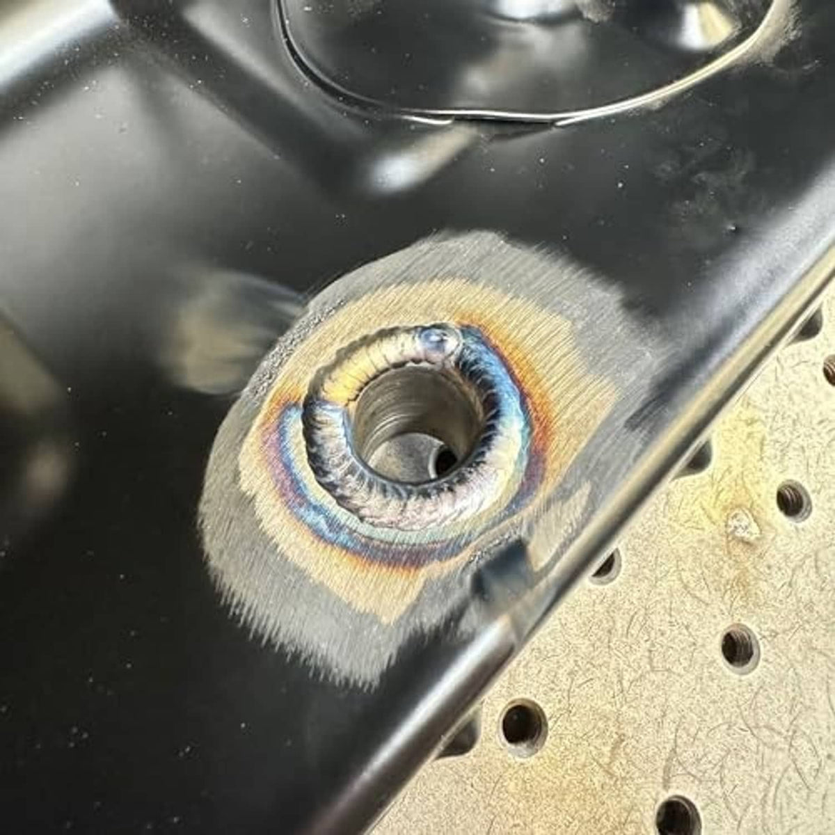Completed weld around male bung fitting on metal surface, heat-tint ring visible