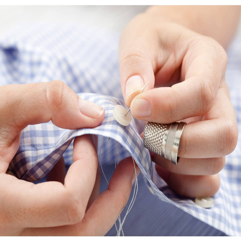 Hands sewing a small round button onto blue check fabric with white thread.