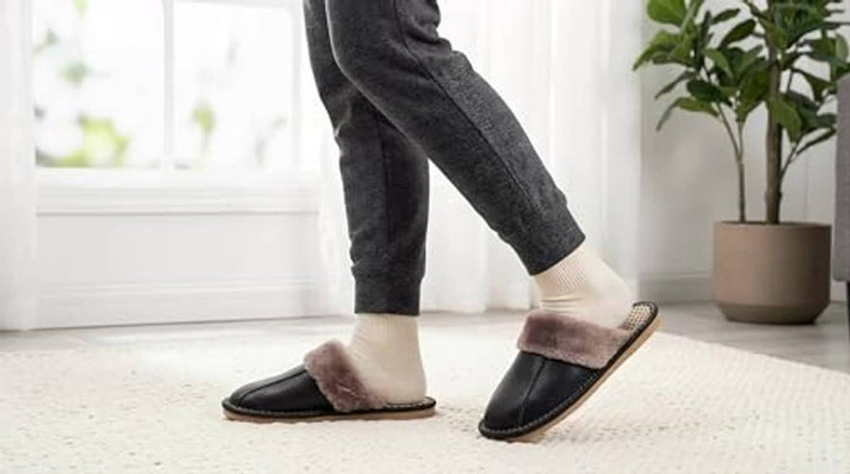 Unisex black leather slip-on slippers with plush taupe lining worn with dark jeans.