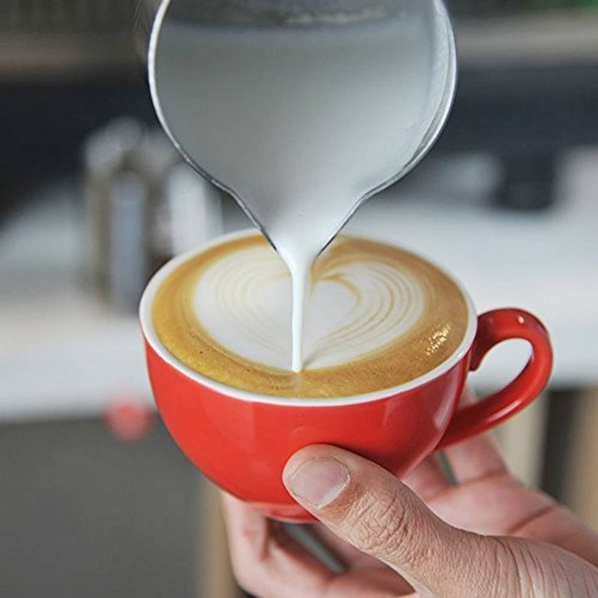 Stainless steel frothing pitcher pouring into a cup, showing thin milk stream