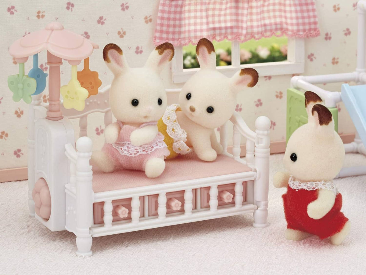 White dollhouse crib with pink bedding and blue bow, with bunny dolls posed beside it