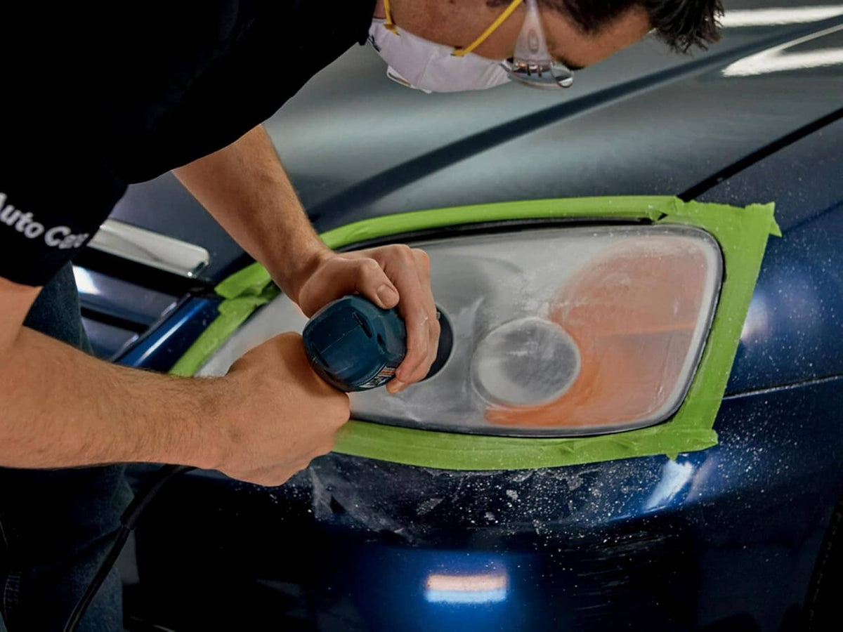 Technician buffs a foggy headlight using a power tool with green masking tape