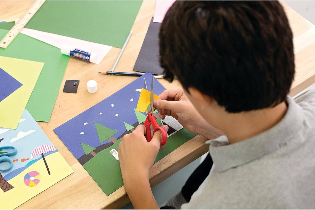 Hands cutting colorful construction paper shapes on a worktable with scissors