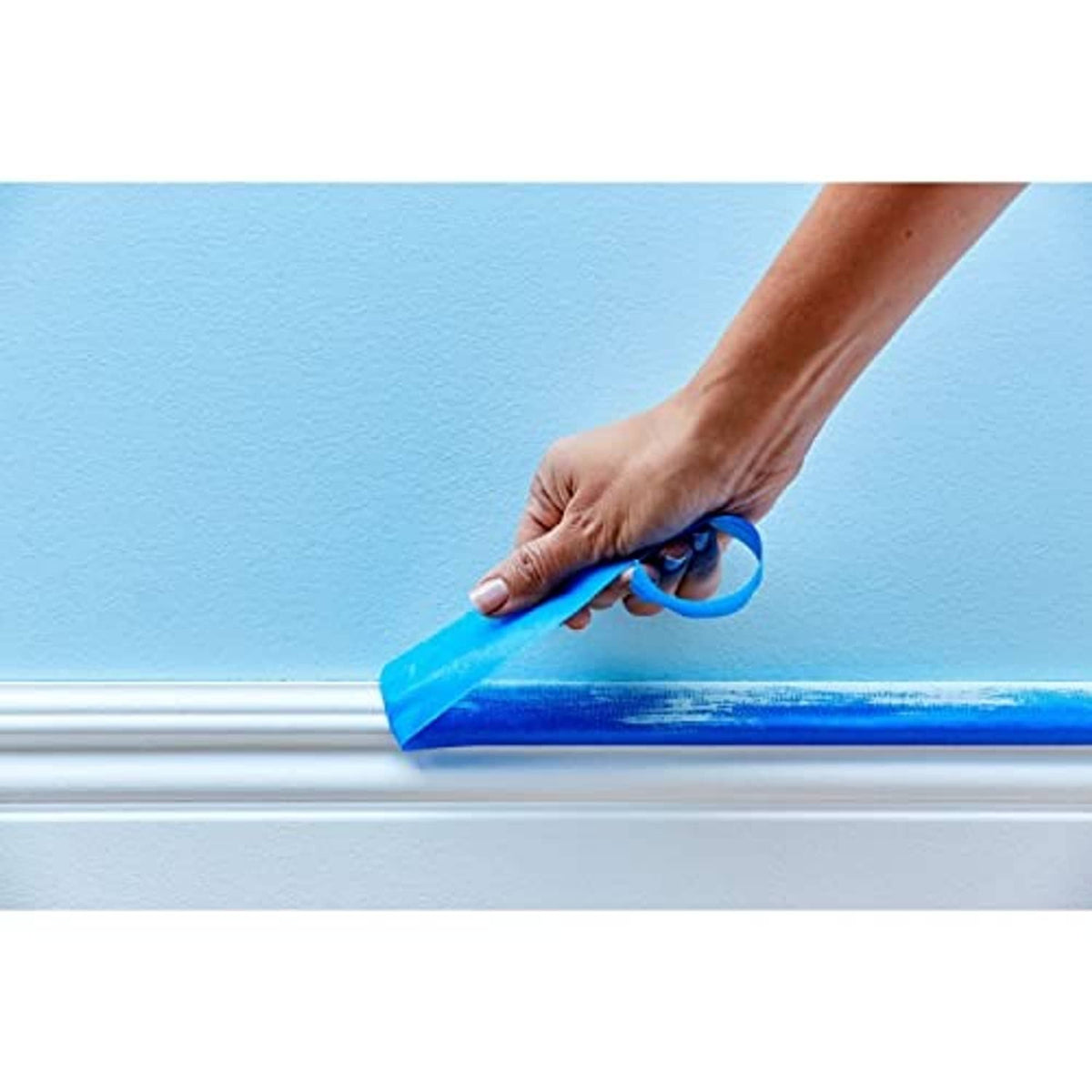 Hand smoothing blue painter's tape along a wall near the base trim