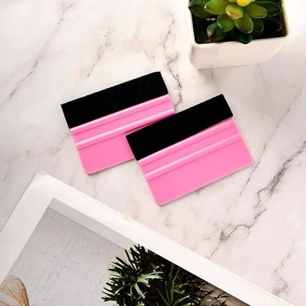 Two pink vinyl squeegee tools with black felt edges laid on a light marble surface
