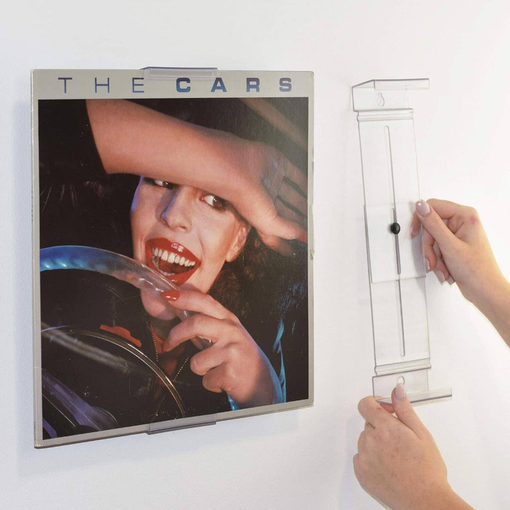 Clear wall-mount album display bracket holding a record cover, hands installing