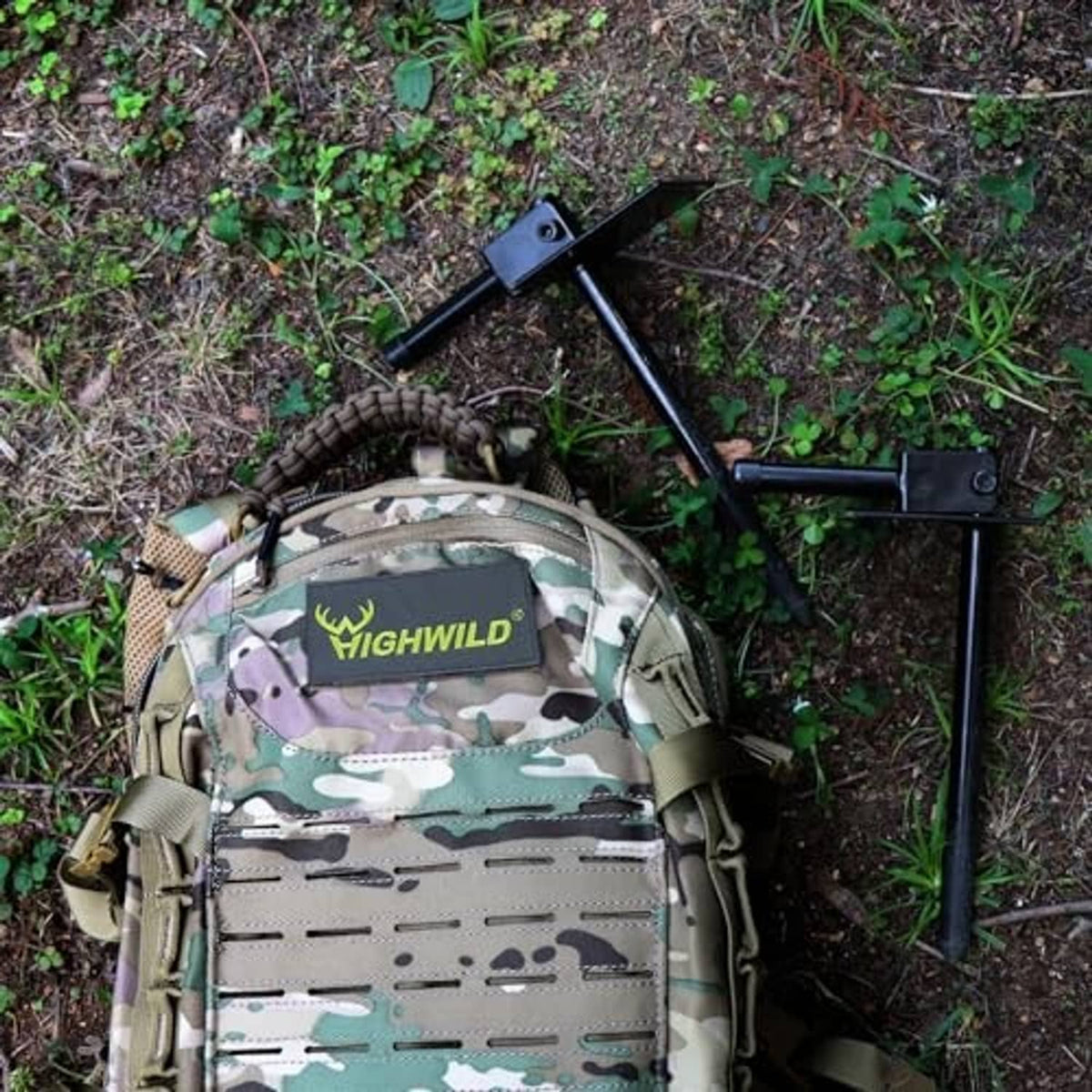 Black ground spike stake and bracket next to a camouflage backpack in outdoor terrain