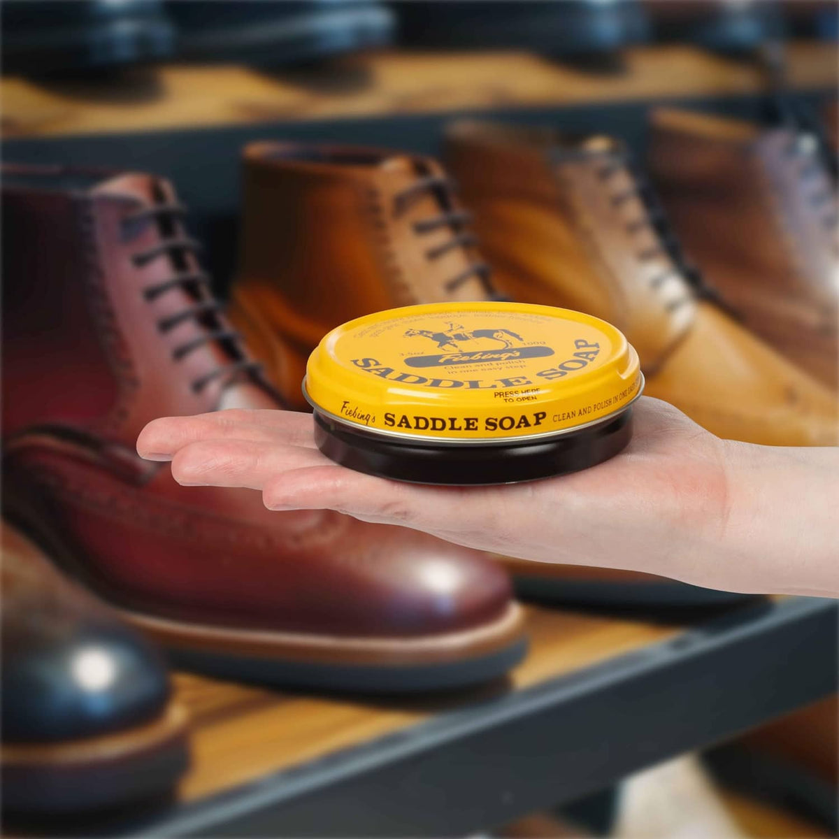 Hand holding a yellow saddle soap tin in front of rows of leather shoes
