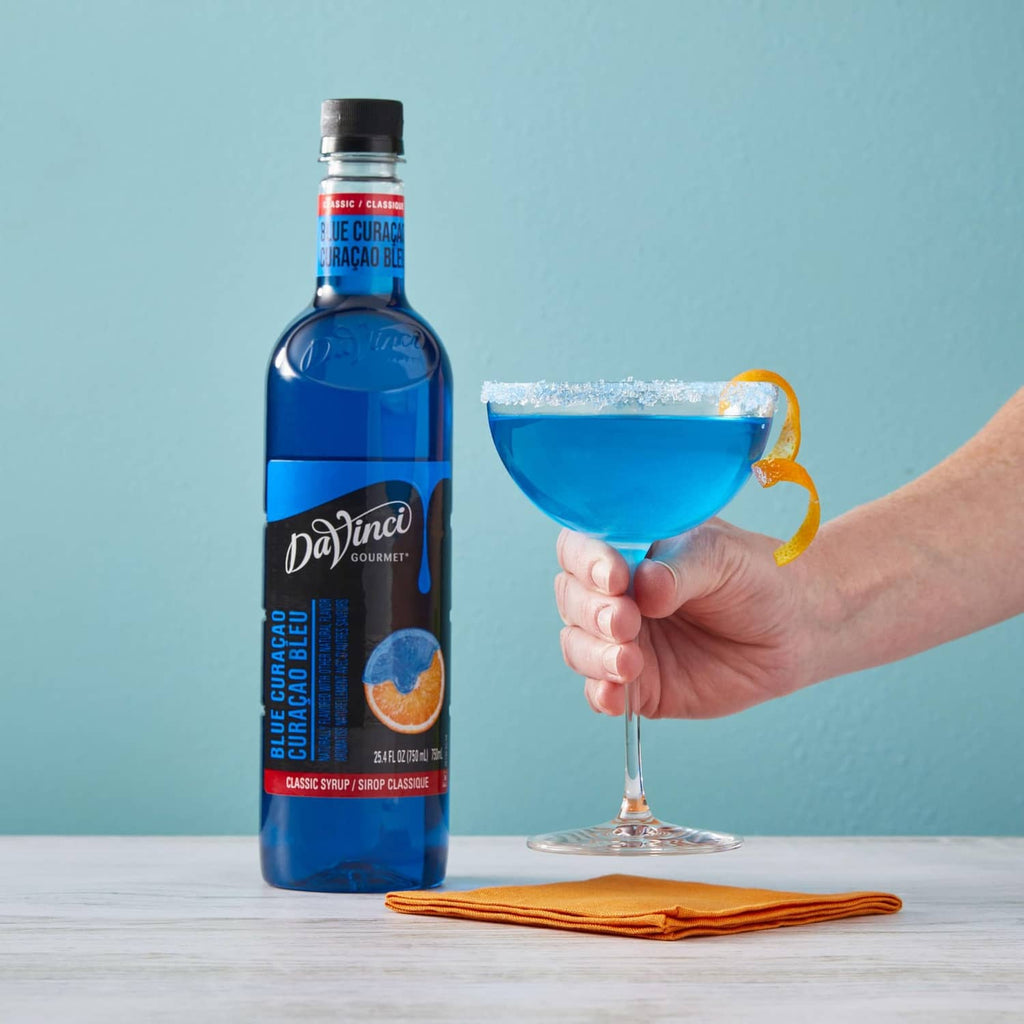 Blue Curacao syrup bottle beside hand holding blue cocktail in a stem glass