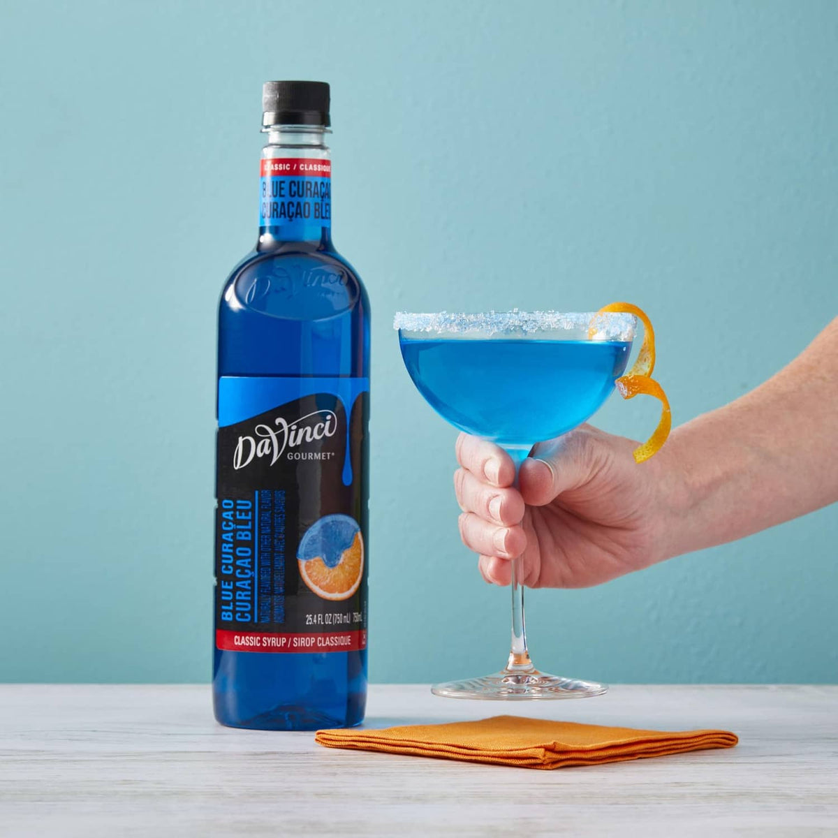 Blue Curacao syrup bottle beside hand holding blue cocktail in a stem glass