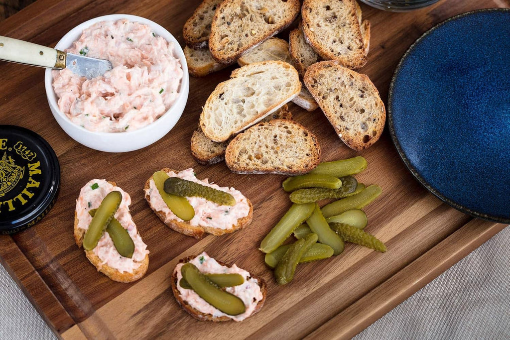Assorted open-faced bread with green cornichons and a bowl of spread on board