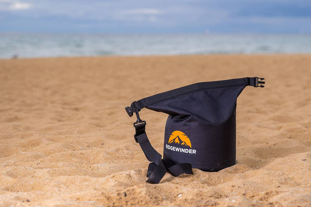 Portable camp shower dry bag with carrying handle and hose on sandy ground
