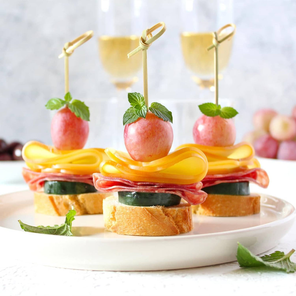Wooden snack picks with fruit and mint on open-faced sandwiches on a plate