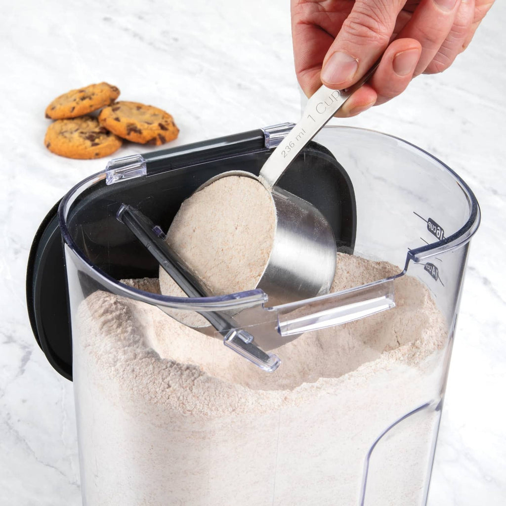 Hand scooping flour from clear airtight container with black lid and utensil holder