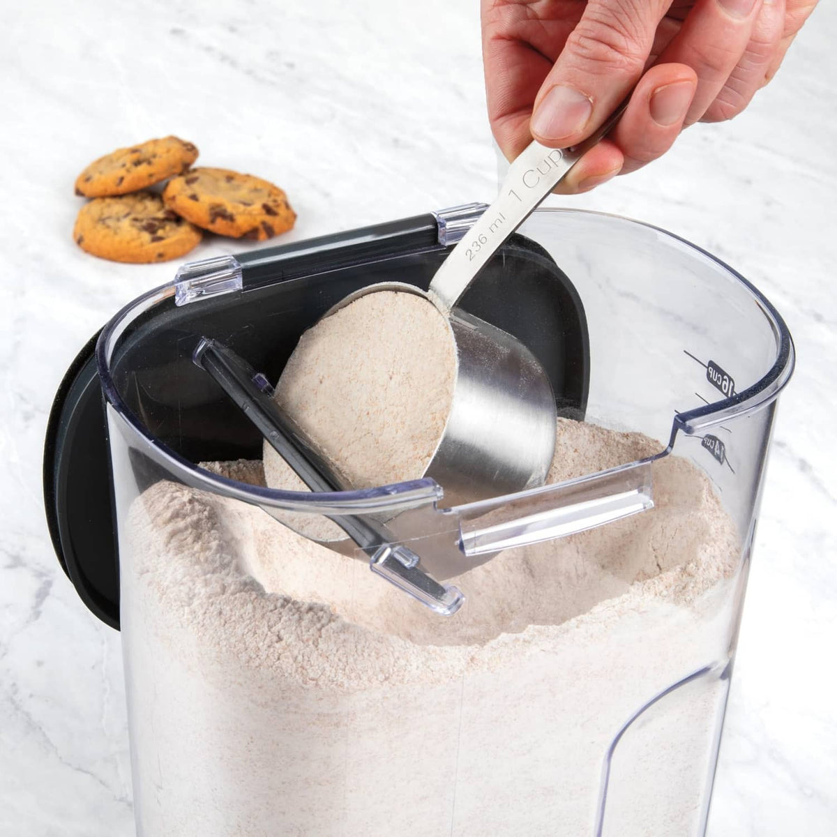 Hand scooping flour from clear airtight container with black lid and utensil holder