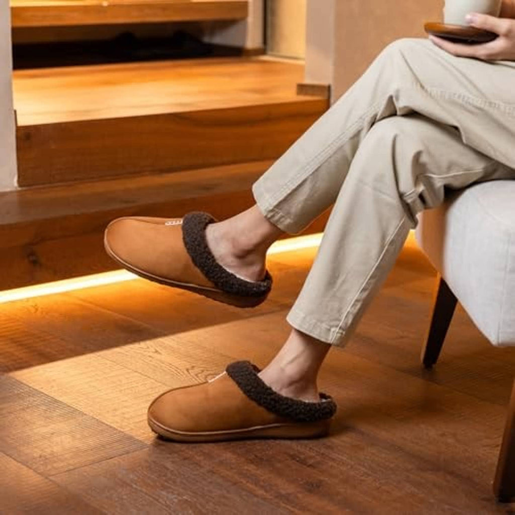 Brown suede slippers with dark plush trim worn with jeans indoors on wood floor