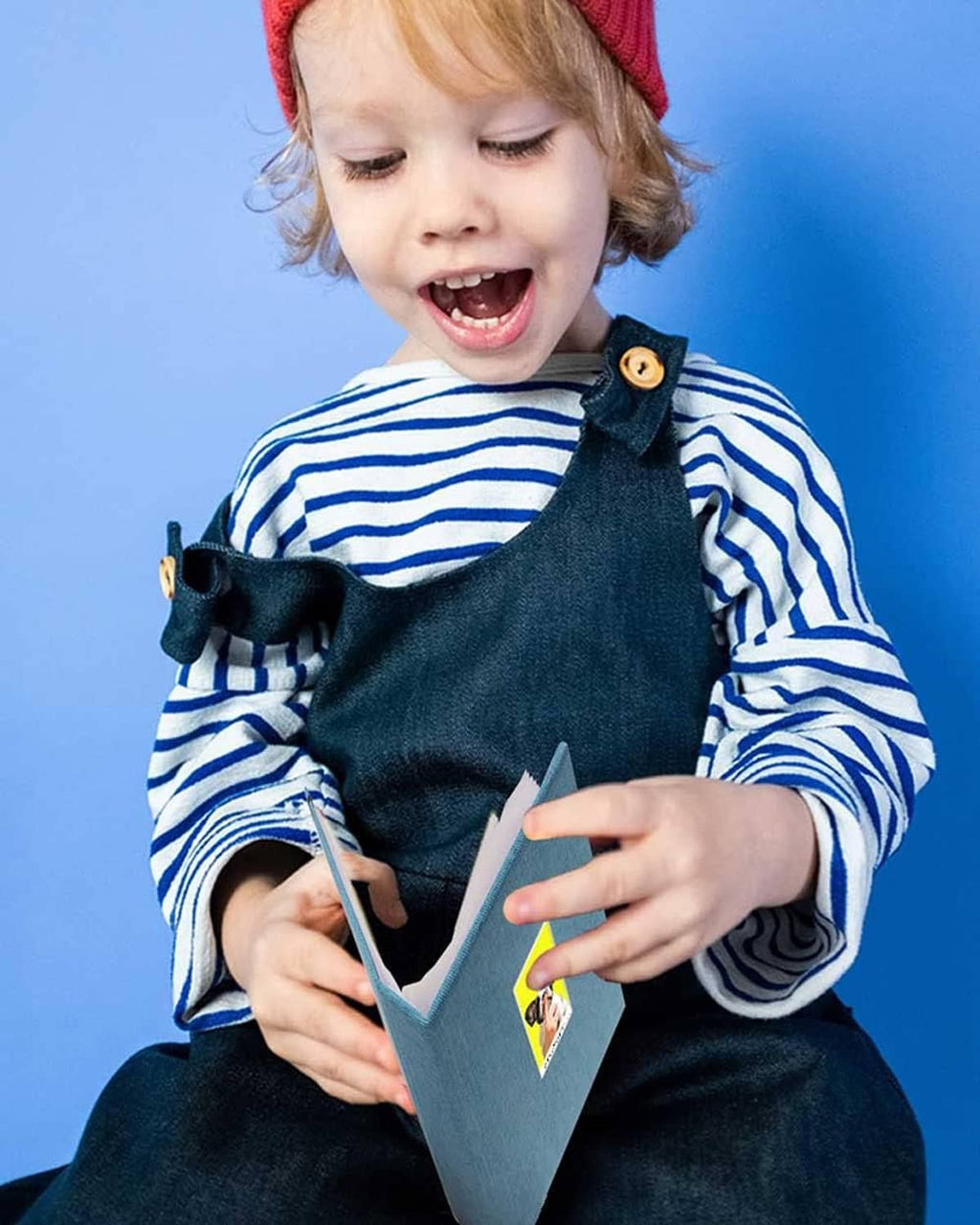 Child holding mini photo sleeves with photos visible; smiling in striped shirt overalls