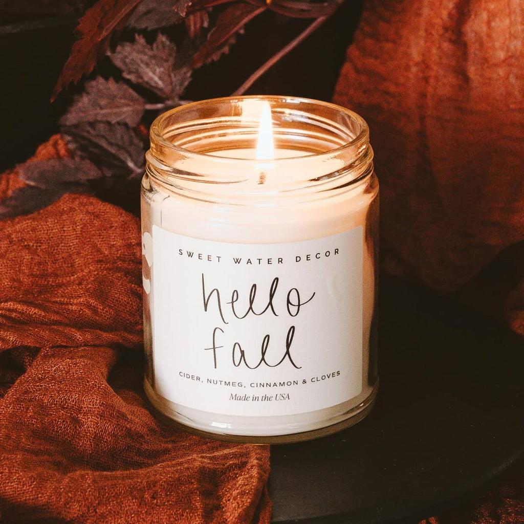 Lit 'Hello Fall' glass jar candle with cinnamon, nutmeg, and clove scent label on white wax