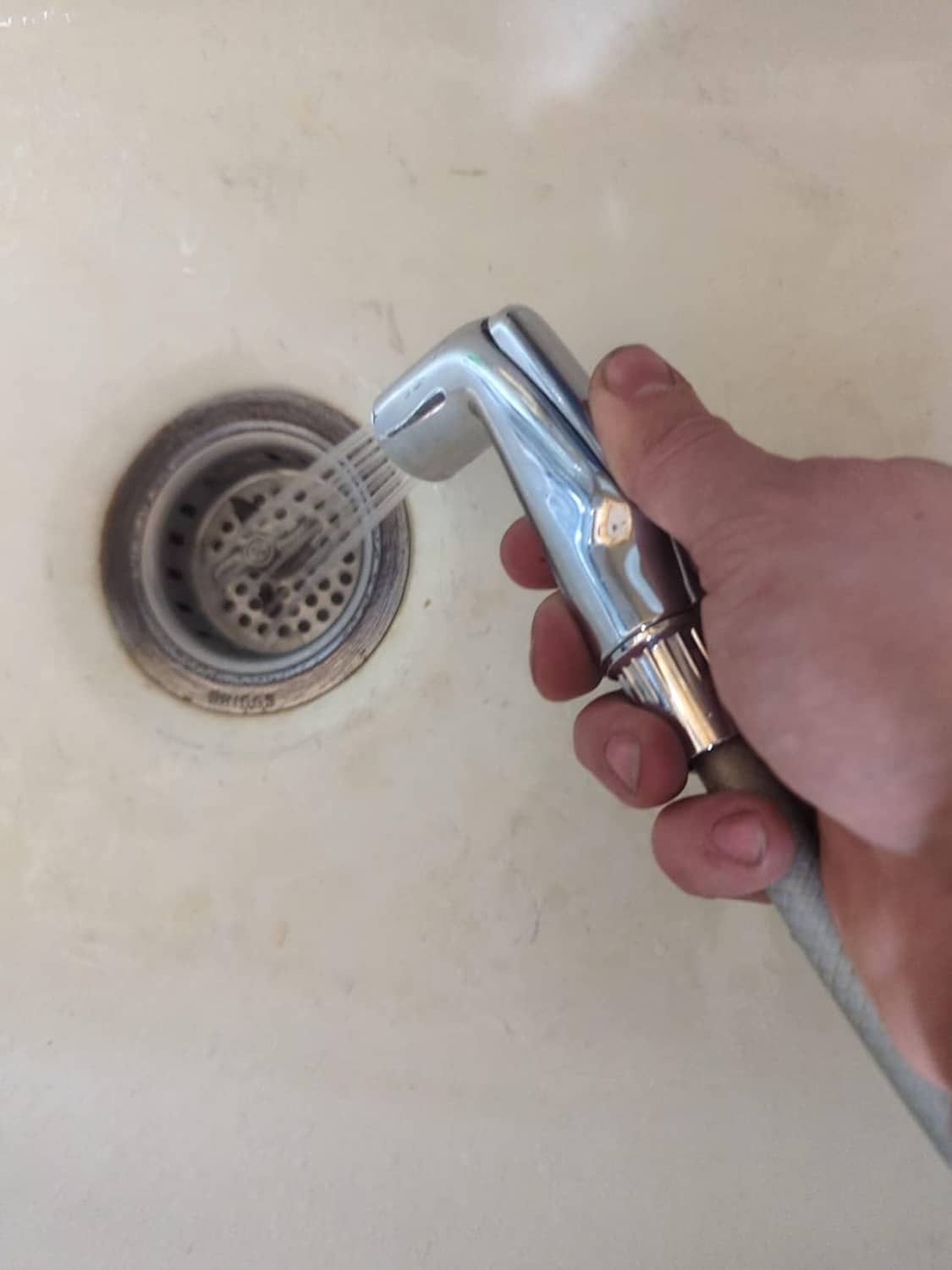 Hand holding chrome spray attachment against sink faucet outlet for cleaning.