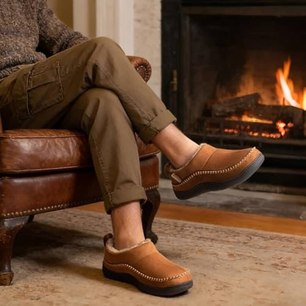 Tan men’s moccasin slippers with faux-fur lining and stitched edge, worn indoors by a fireplace.