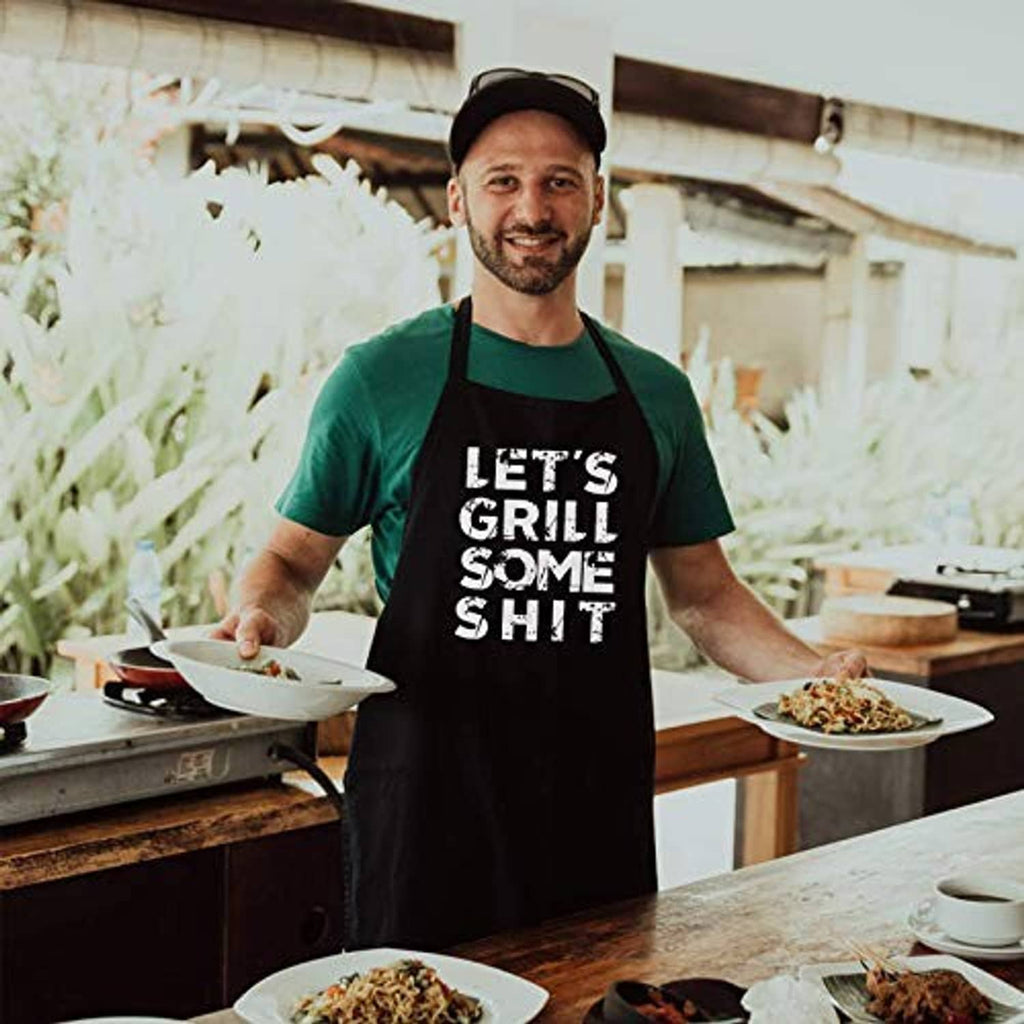 Black BBQ apron with "LET'S GRILL SOME SHIT" worn by man in green shirt outdoors