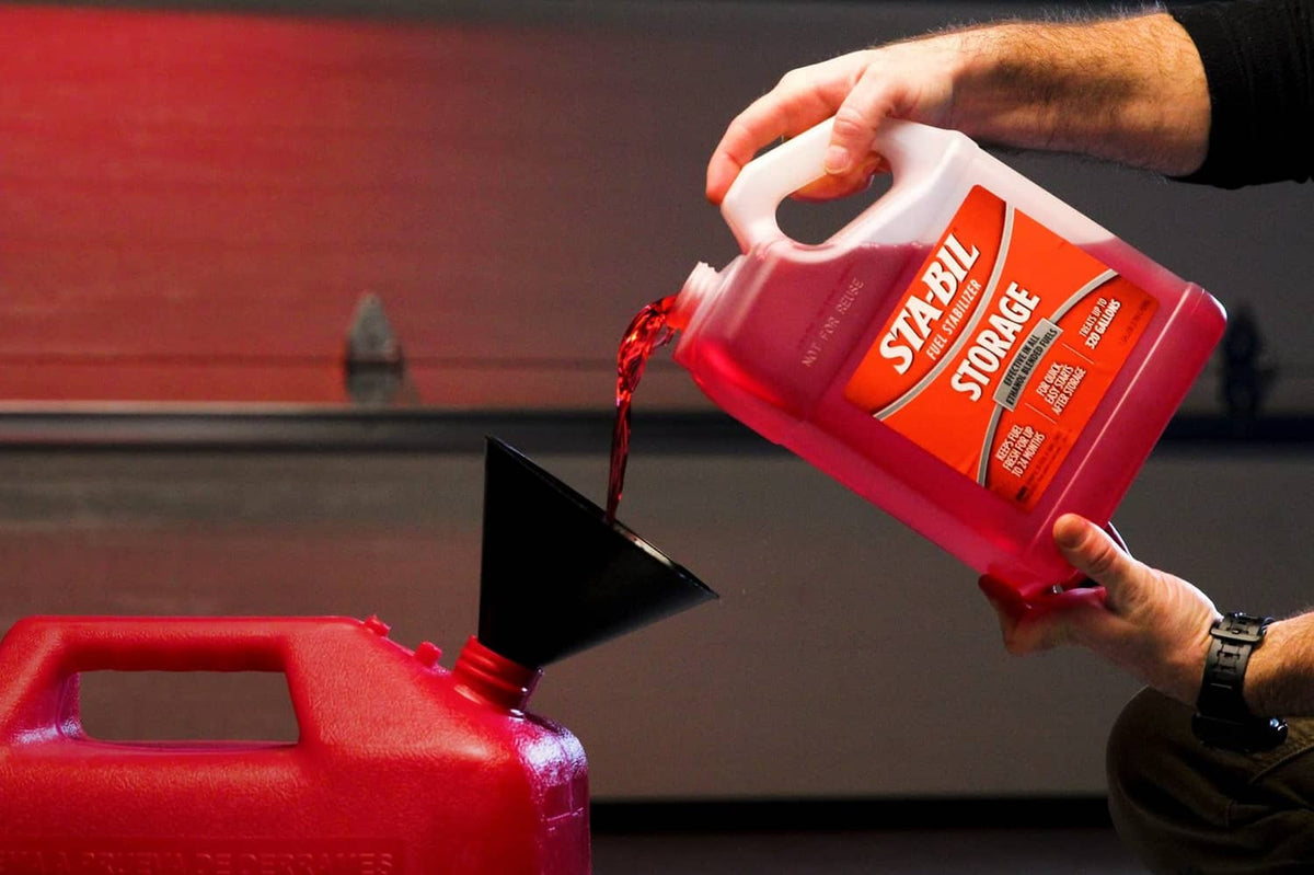 Tilted red fuel stabilizer jug pouring into red fuel container with stream visible