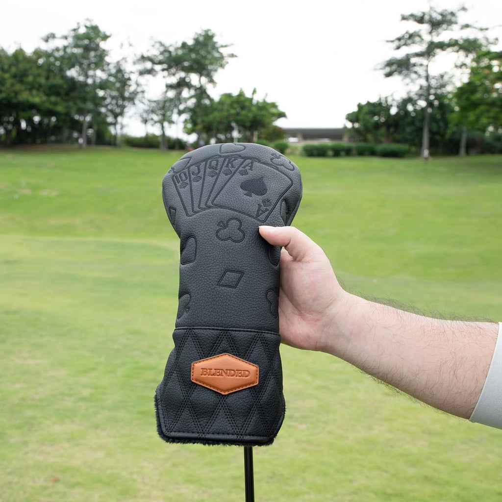Black driver golf club head cover held by hand, showing tan logo patch and poker pattern.
