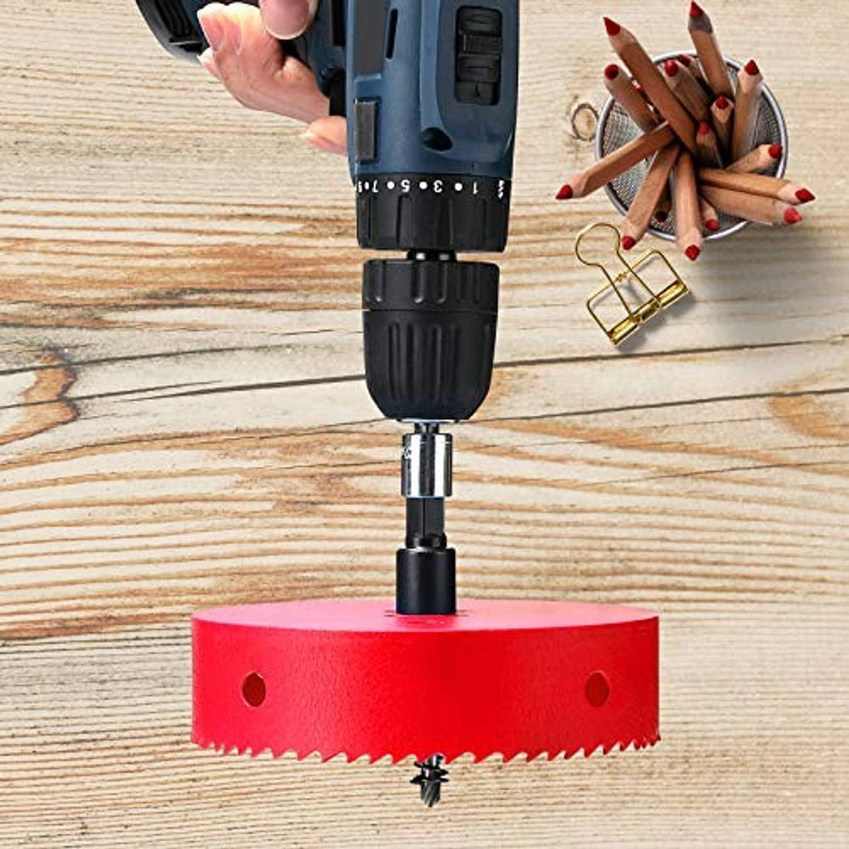 Red bi-metal hole saw mounted on a drill, cutting into a wooden board.