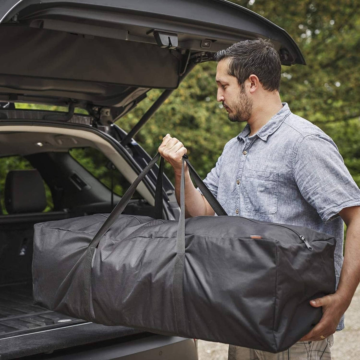 Black long duffel bag loaded into open car trunk, held by carrying handles