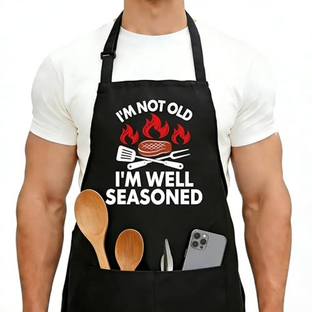 Black cooking apron front view with flaming grill icon, utensils, and “I’m well seasoned” text