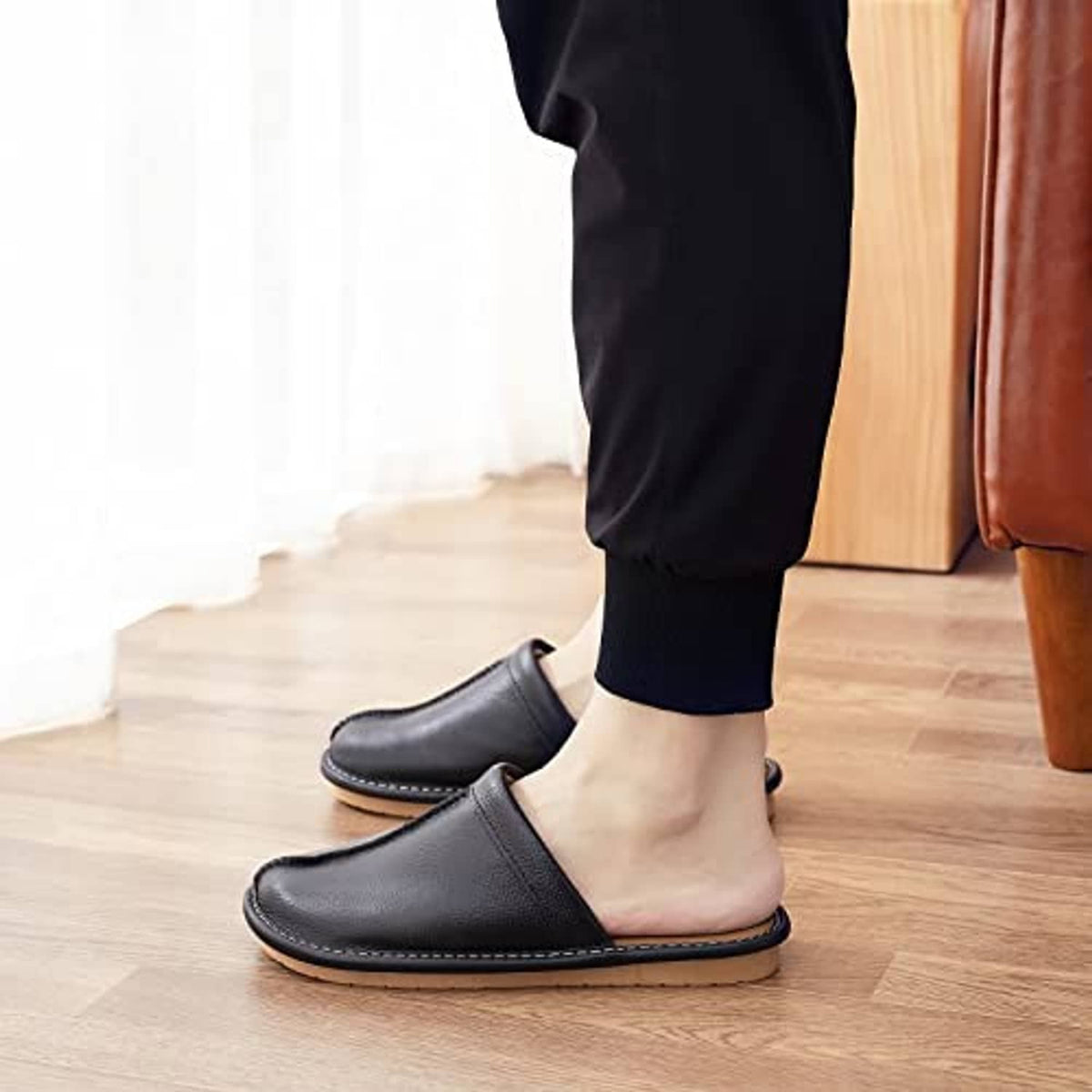 Black leather slip-on slippers worn indoors with low-profile sole on light wood