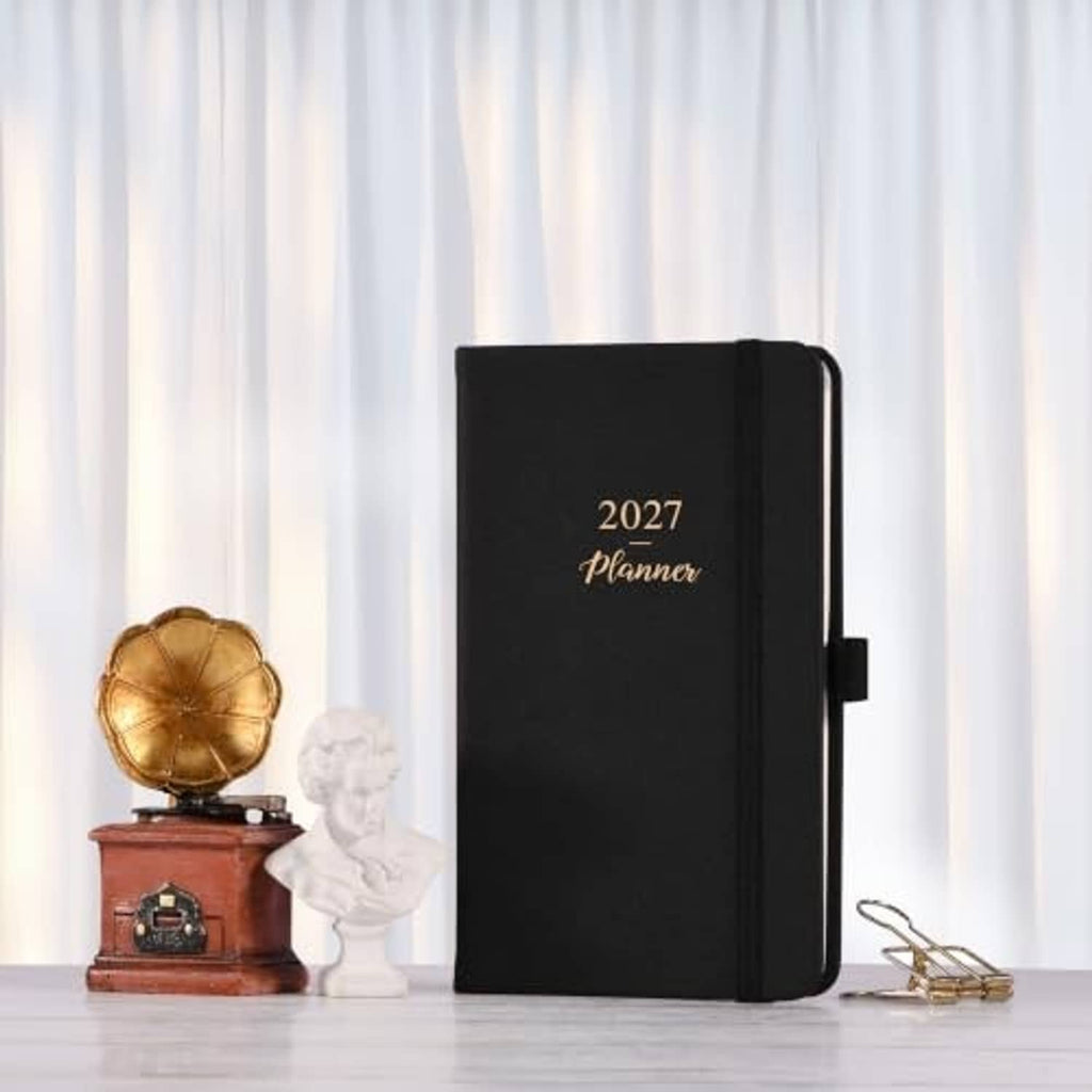 Black 2027 pocket planner on desk with ribbon-style band and carry handle on the side