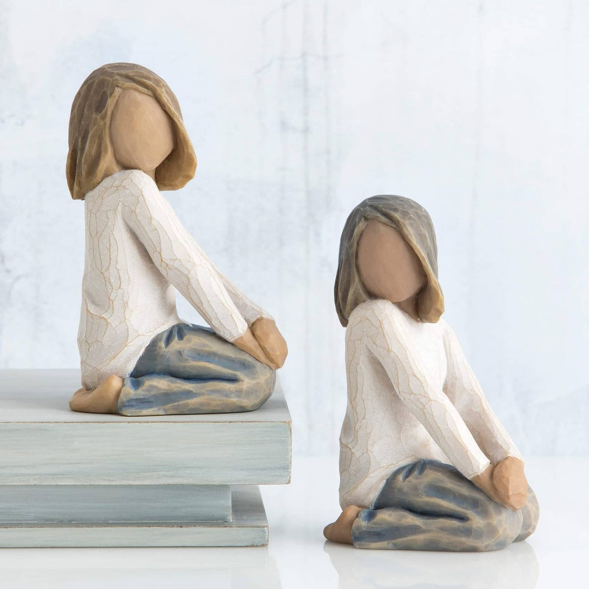 Two kneeling child figurines with brown hair and white robes, blue-gray blankets, on bench.