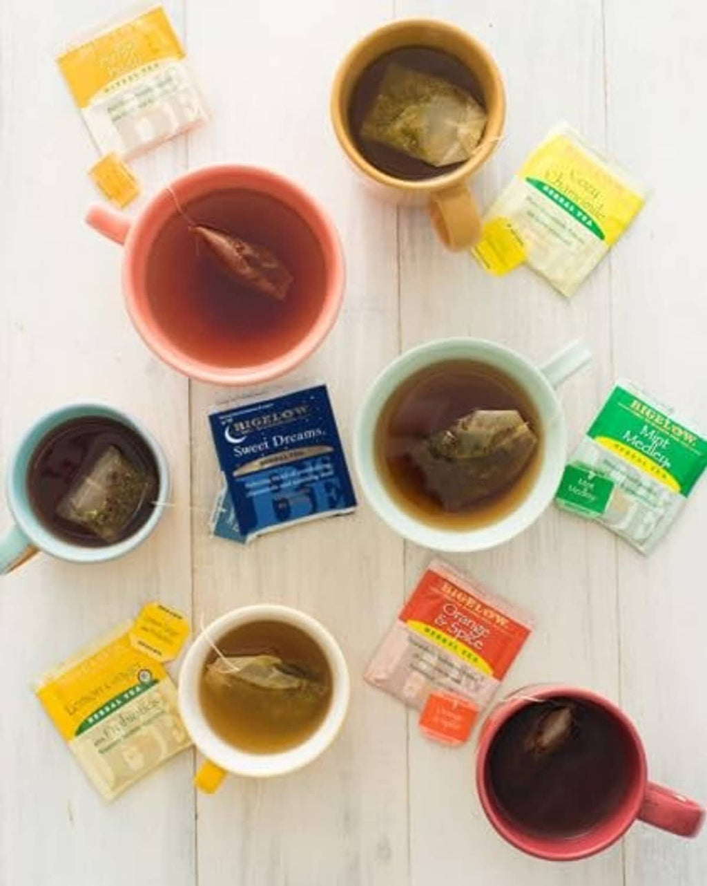 Assorted herbal teas in colored mugs with tea bags, multiple cups on a wood surface