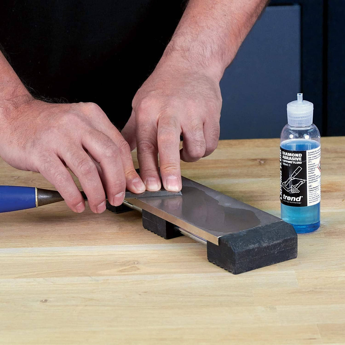 Hands lapping a metal blade while a blue diamond abrasive fluid bottle stands nearby