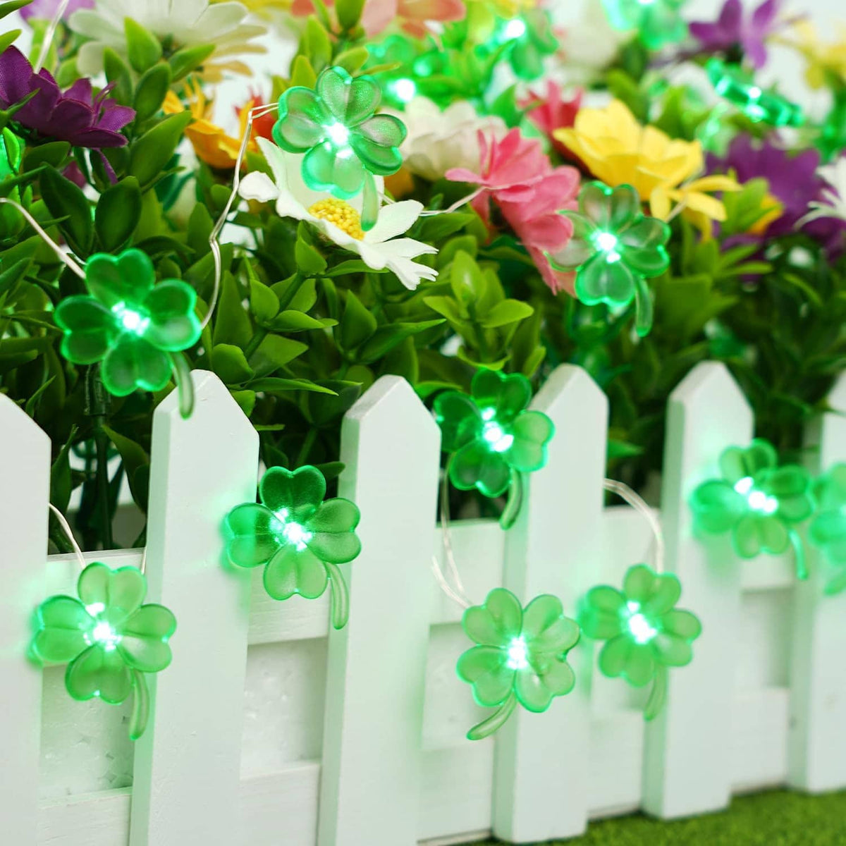 Green glowing shamrock string lights with clover-shaped LEDs on outdoor flowers and white picket fence
