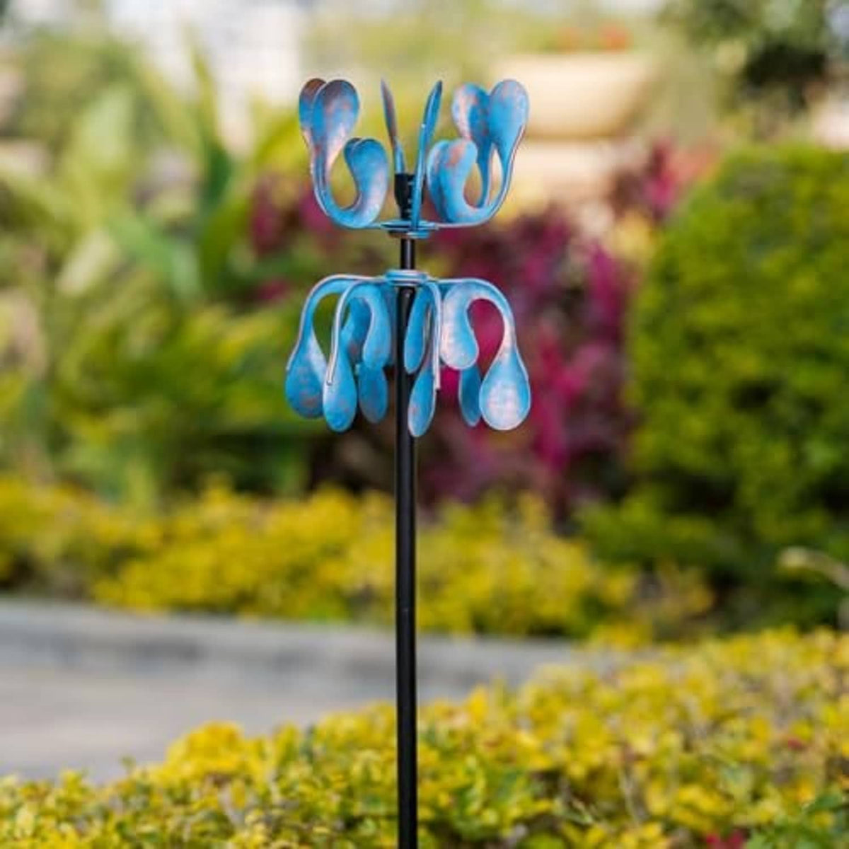 Blue metal wind spinner with curled petal blades on black stake in garden