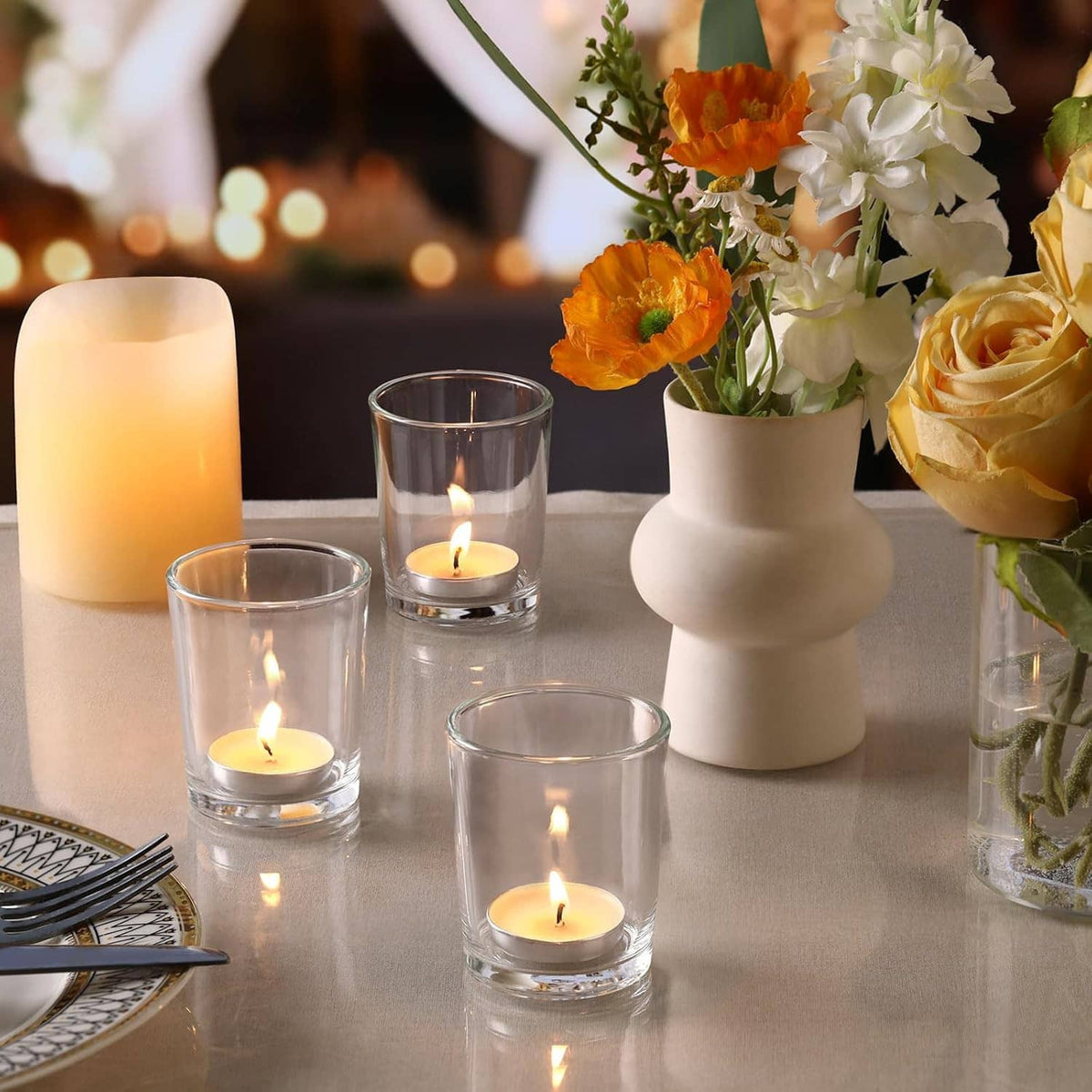 Three clear glass tea light holders with lit candles beside flowers on a table