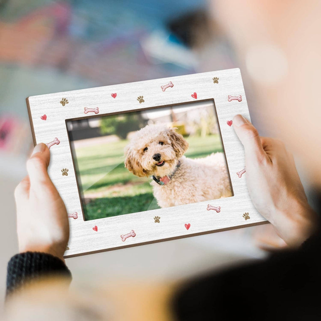 White dog photo frame with paw and bone pattern, held in hands over dog portrait.