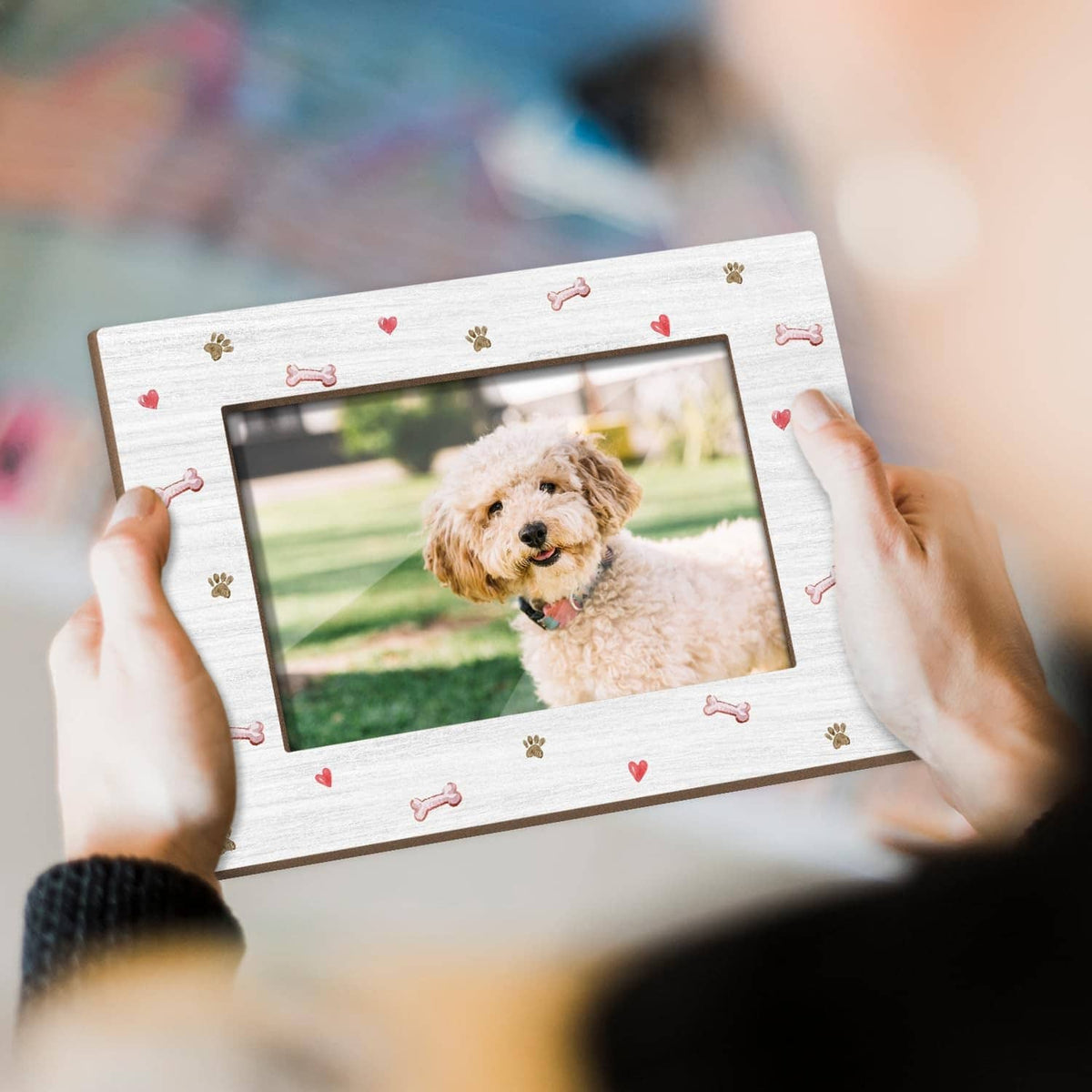 White dog photo frame with paw and bone pattern, held in hands over dog portrait.