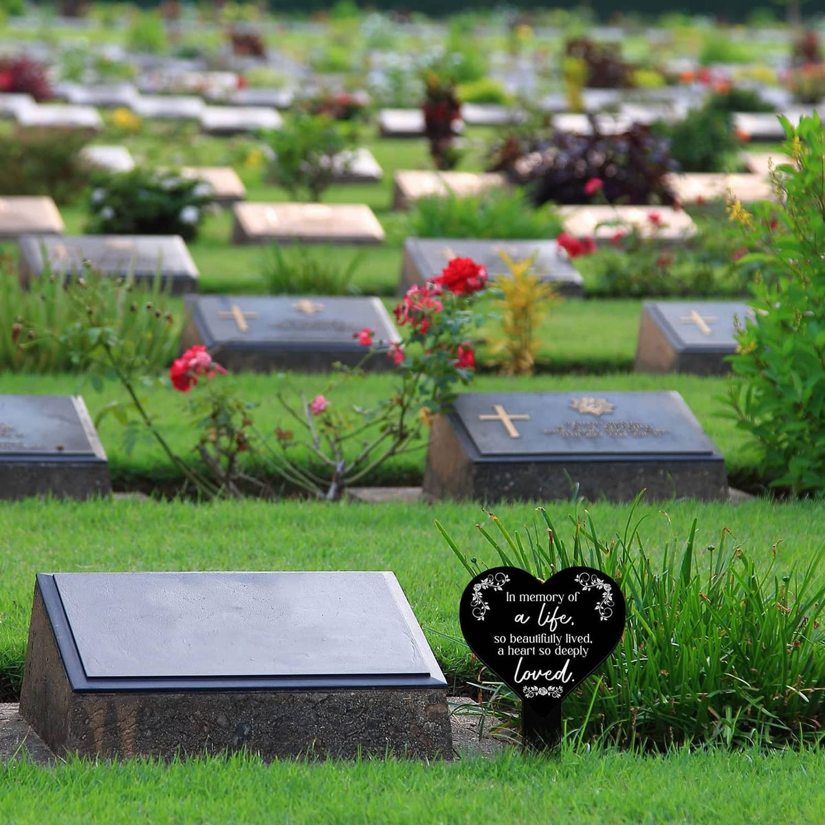 Heart-shaped black remembrance plaque with white verse on stake in a cemetery bed.