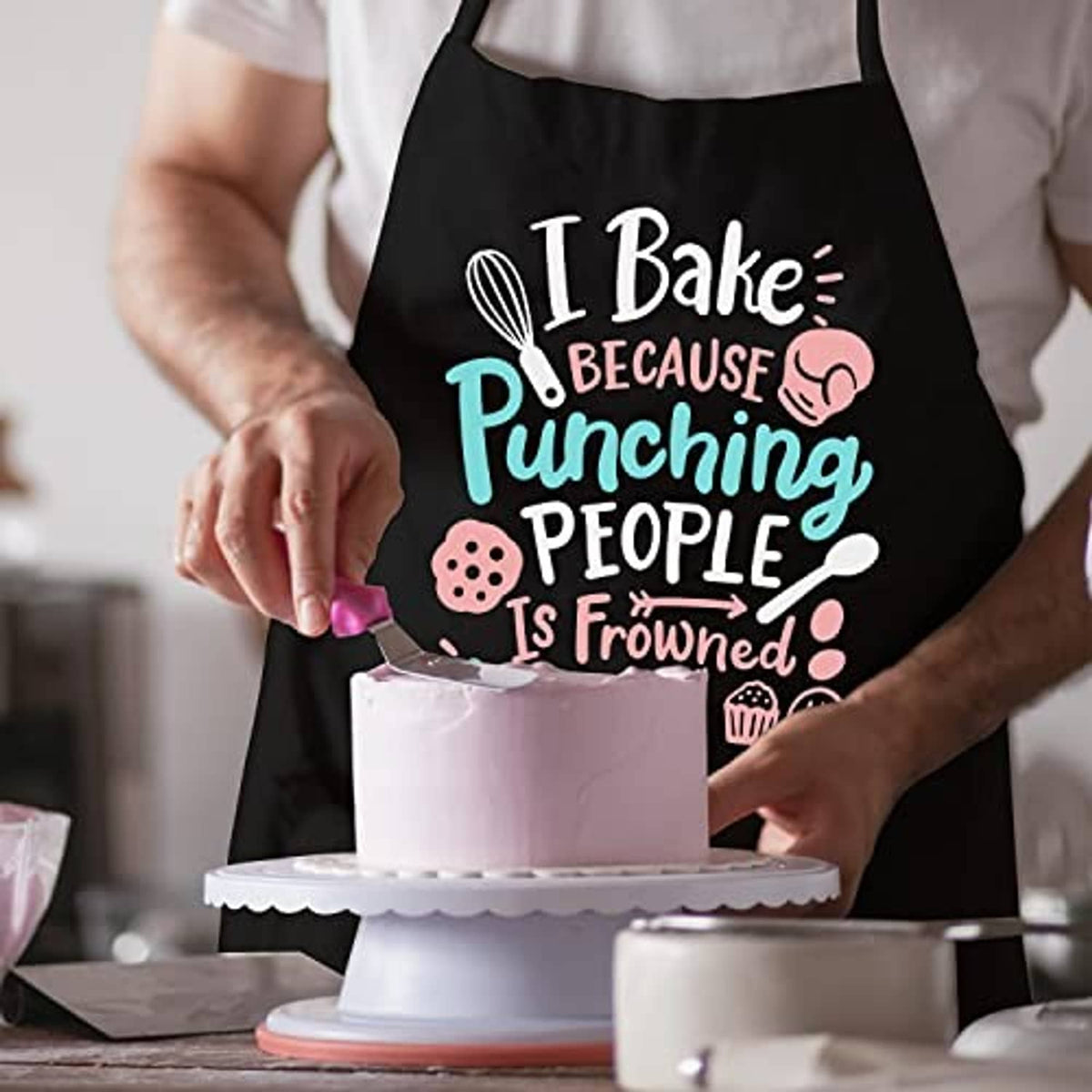 Black baking apron with 'I Bake' slogan while frosting a cake on a stand