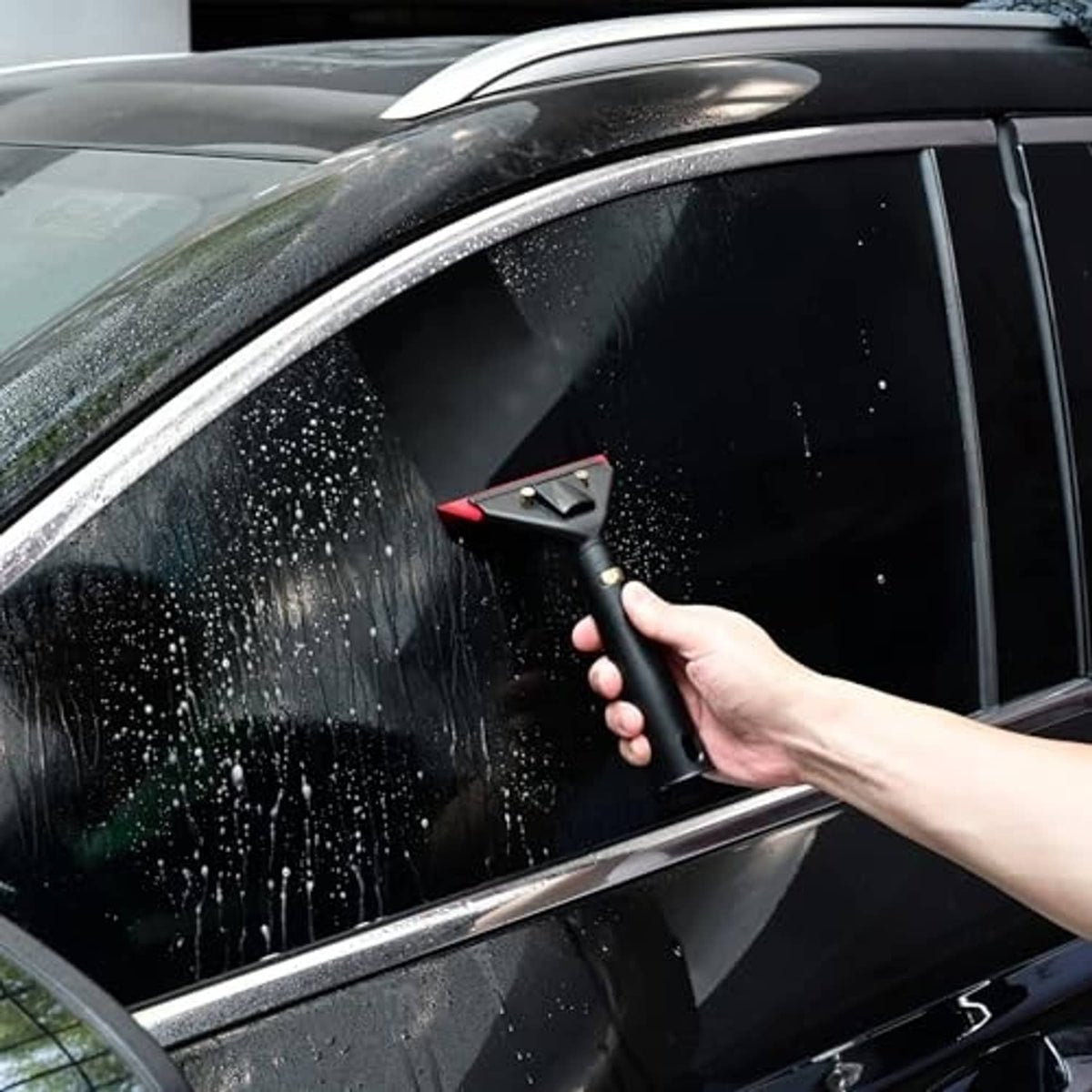 Hand uses small squeegee on wet car window glass with red-edged squeegee head