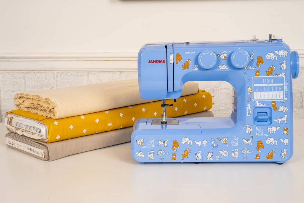 Blue Janome sewing machine beside folded fabric rolls, cat decals visible on body