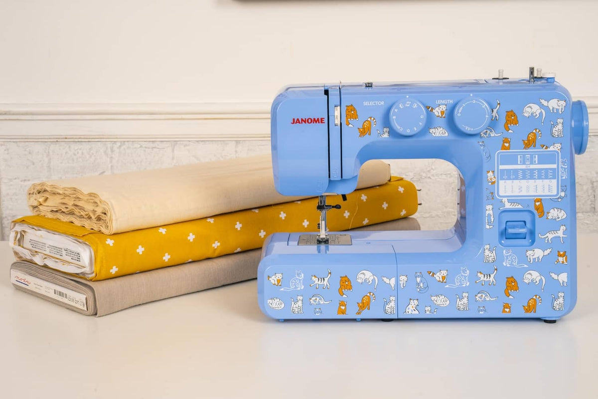 Blue Janome sewing machine beside folded fabric rolls, cat decals visible on body