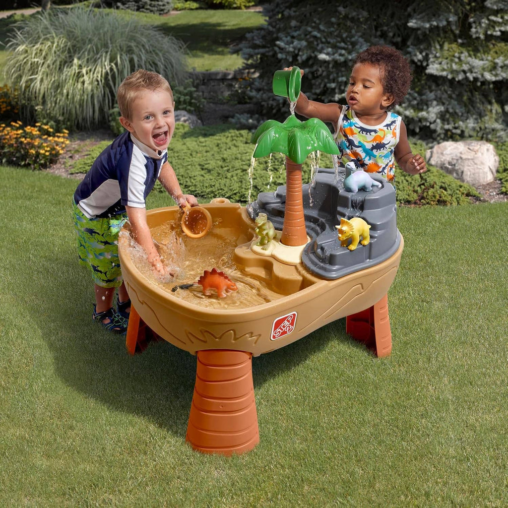Kids dinosaur sand and water play table in lawn with green palm, tan basin, grey water feature