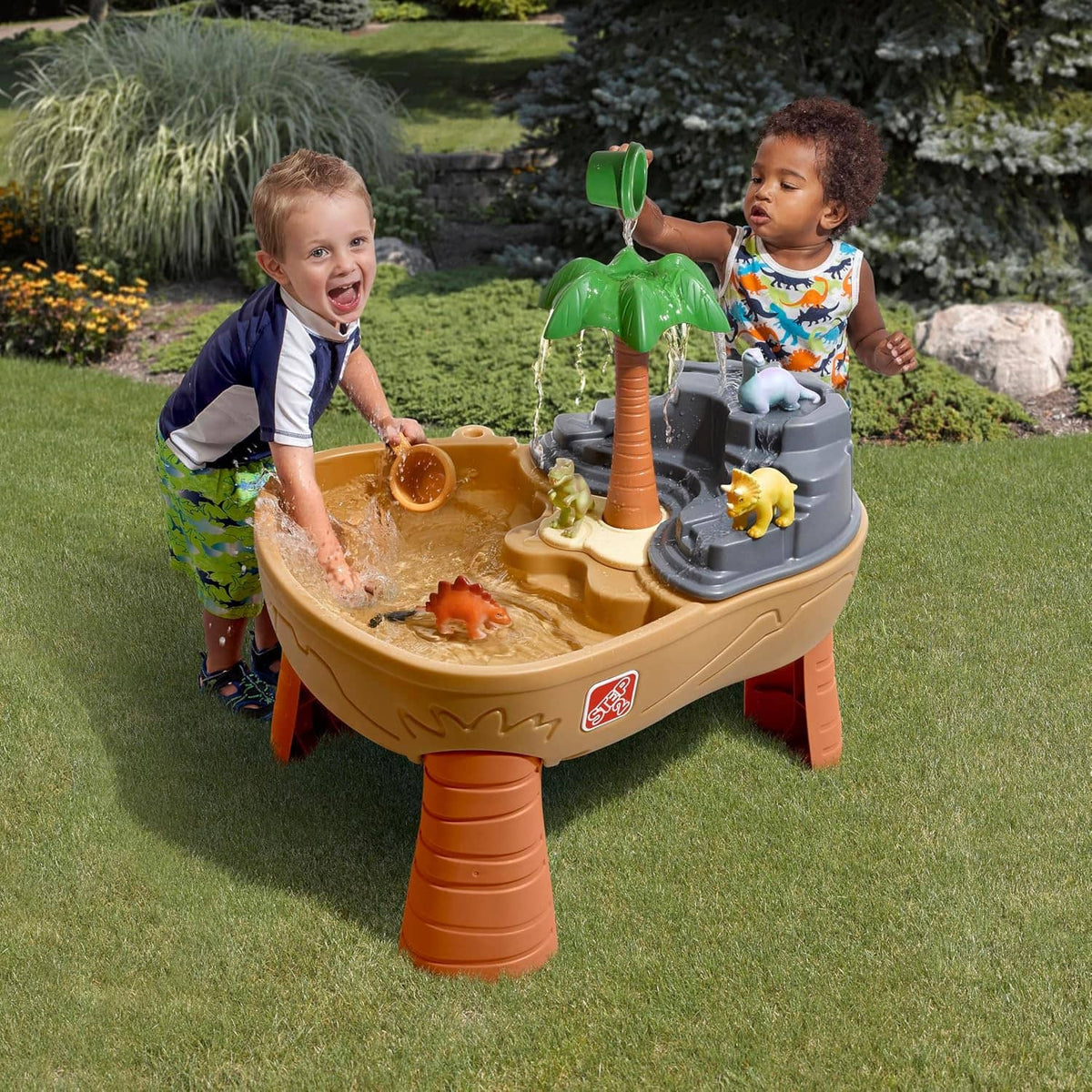 Kids dinosaur sand and water play table in lawn with green palm, tan basin, grey water feature