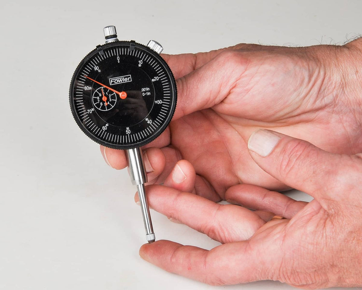 Hand holding a dial indicator gauge with black dial and orange pointer